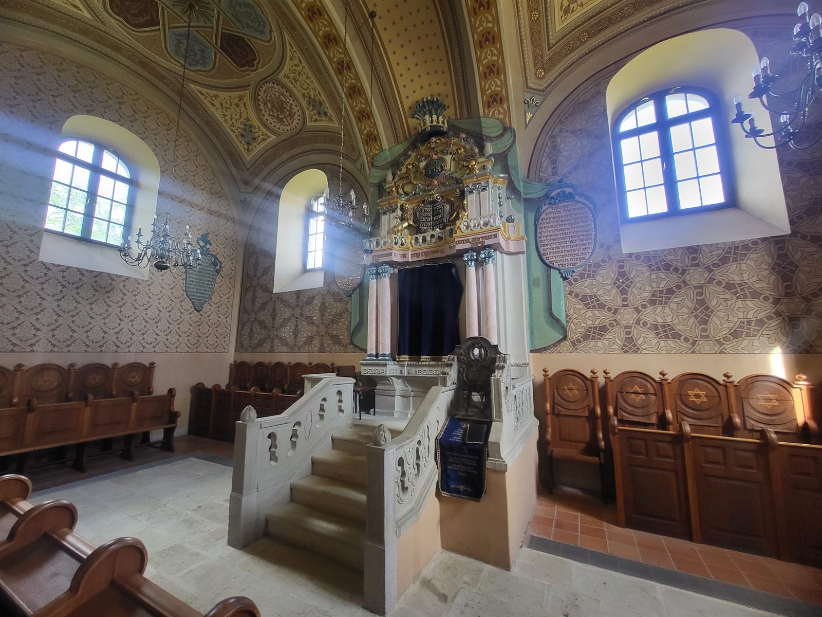 Interior of the Mád synagogue: the ornate Torah ark with marbled columns and gilded crown, the raised bimah with carved stone balustrade, wooden benches and painted walls