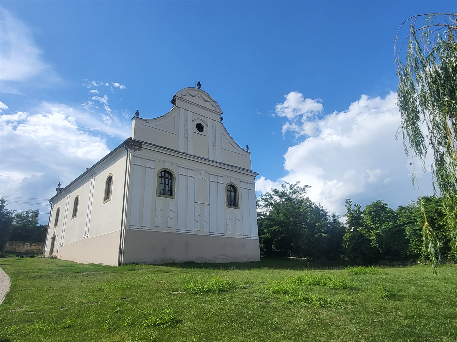 The late-Baroque synagogue of Mád, a white building with a curved gable, standing on a grassy slope under a blue sky