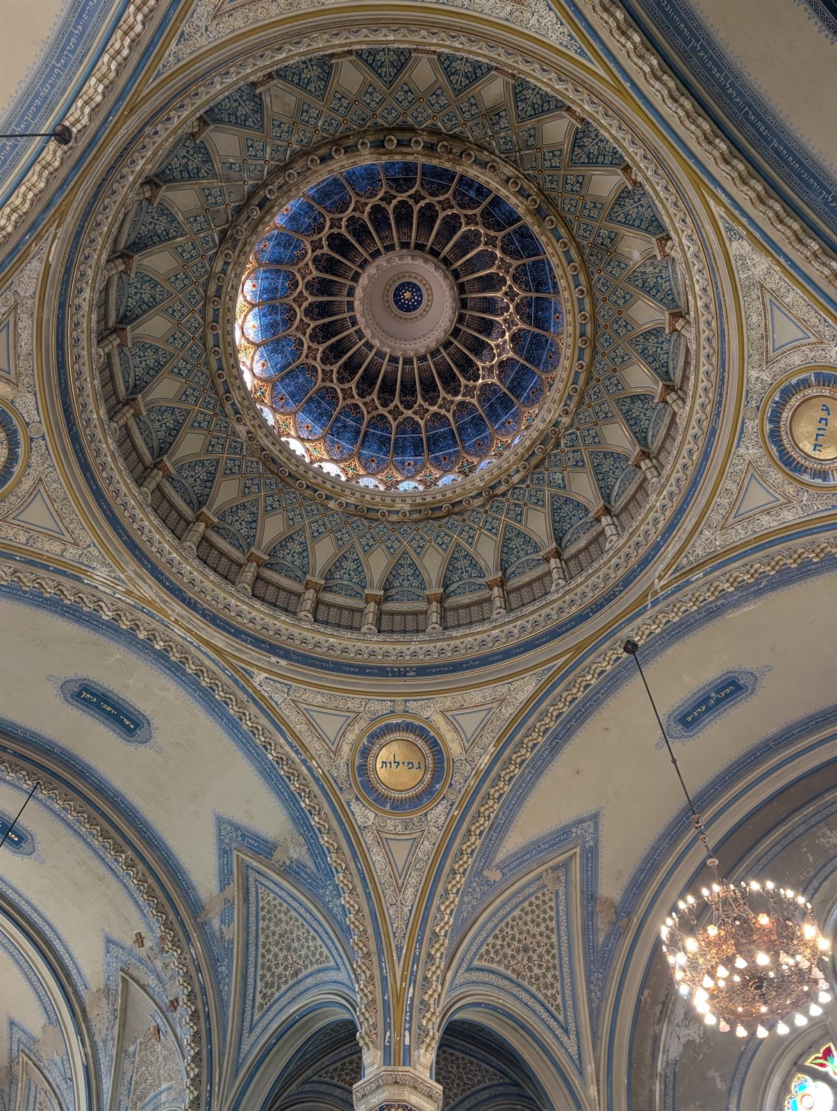 Looking straight up into the dome of the Szeged synagogue: a ring of blue stained glass with white flowers at the centre, surrounded by tiers of painted arches, ornament and gilded medallions