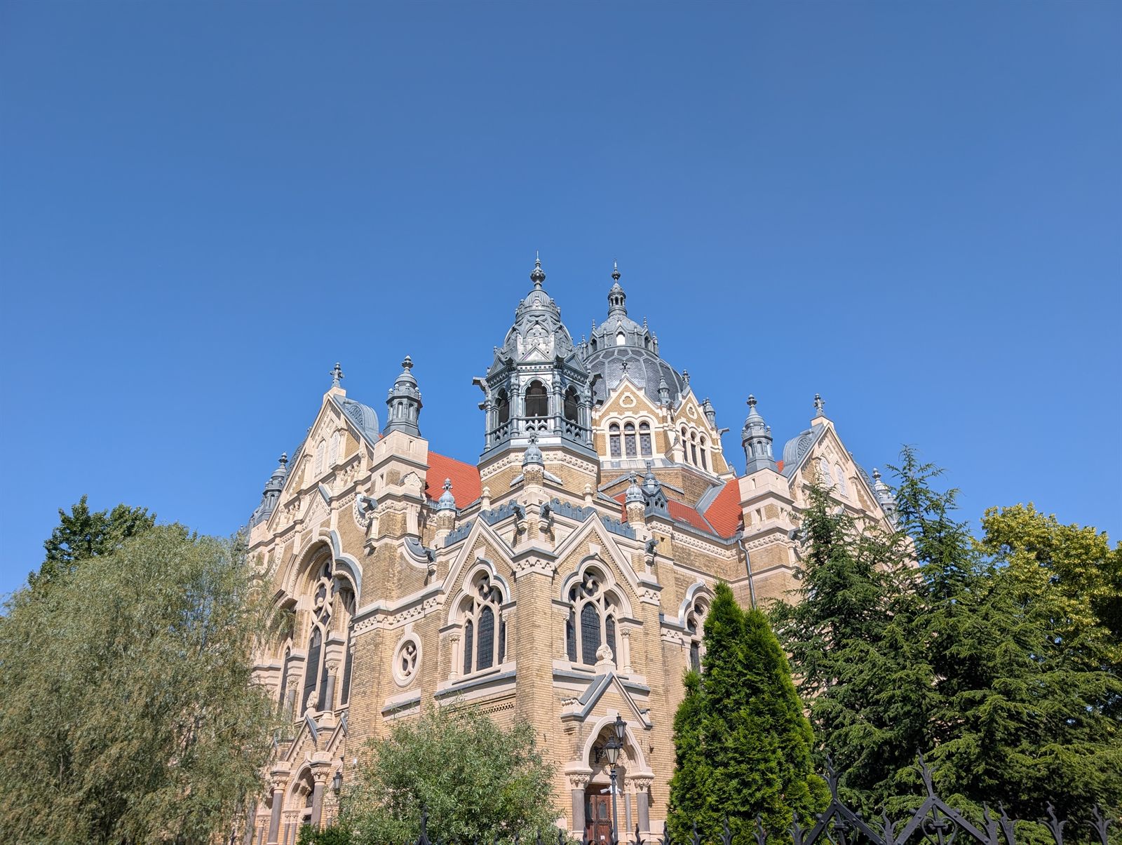 The New Synagogue in Szeged seen from the street: a brick and stone facade with pointed gables, corner turrets and a great central dome against a clear blue sky