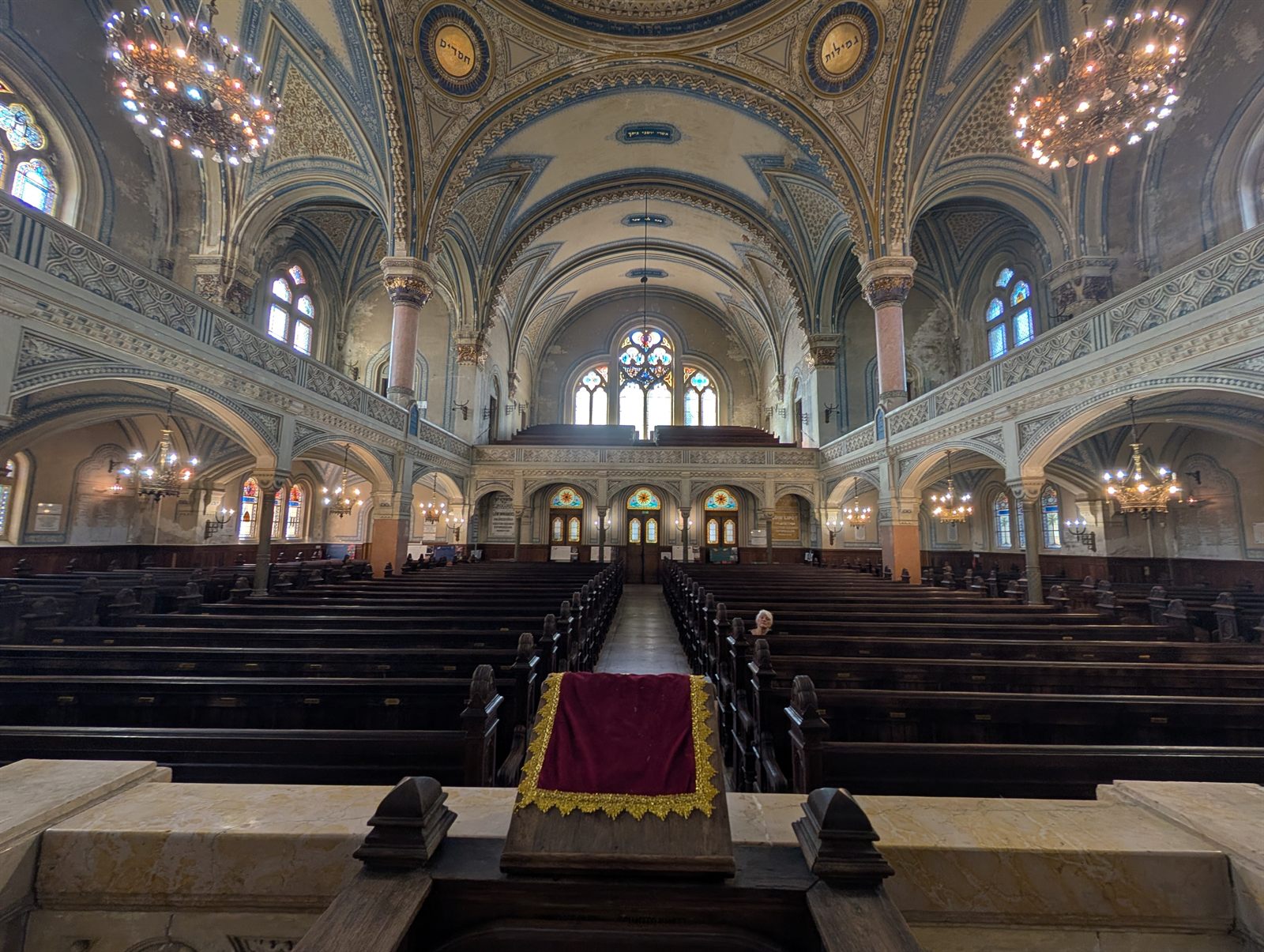 The sanctuary of the Szeged synagogue seen from the bimah: rows of dark wooden benches under painted vaults, arcaded galleries on both sides, chandeliers and the organ loft at the far end