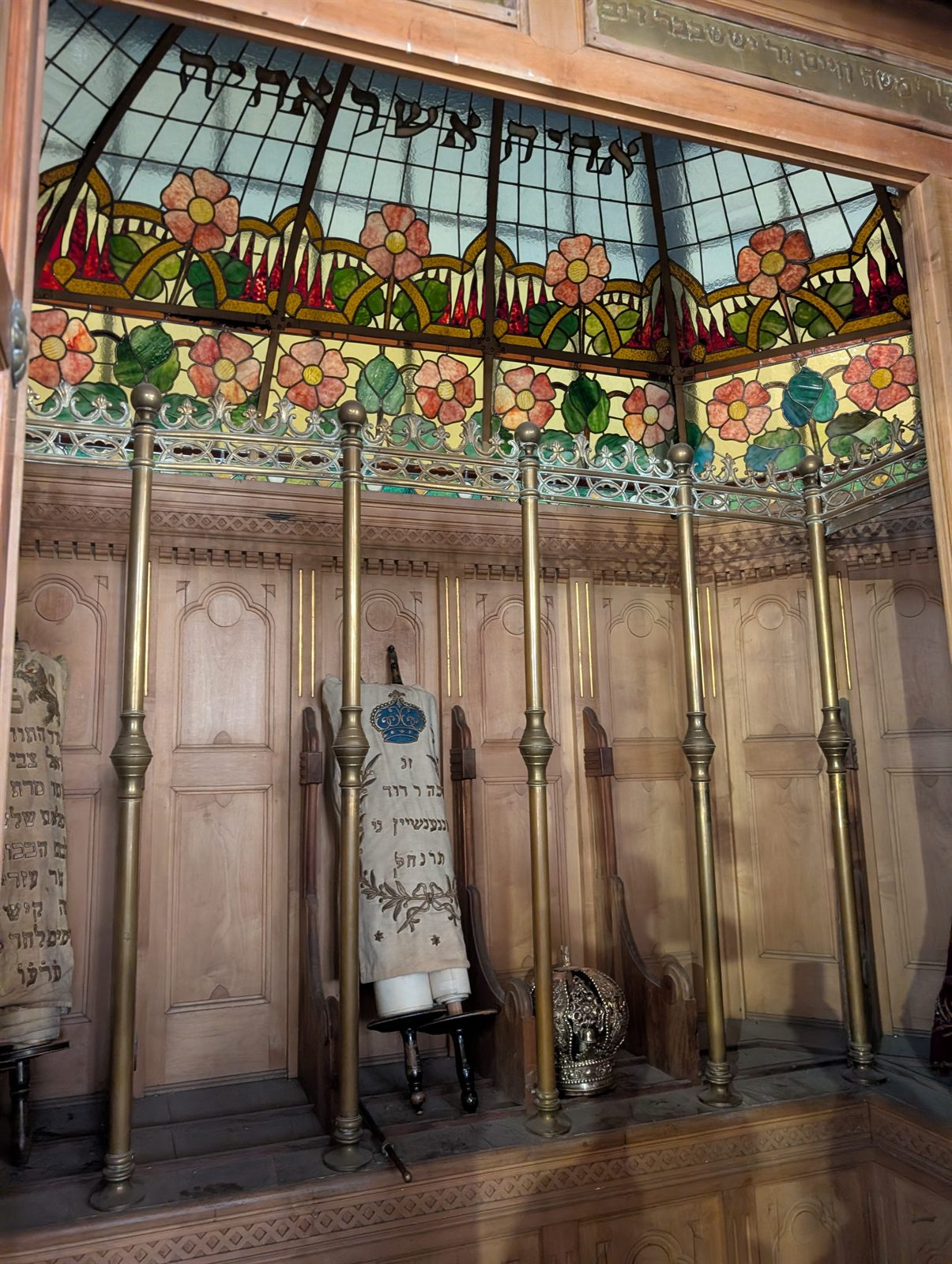 Inside the open Torah ark at Szeged: Torah scrolls in white mantles standing behind brass rods, under a stained-glass canopy of pink flowers and green leaves