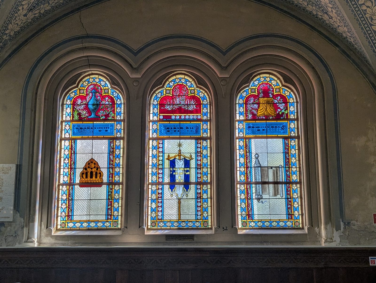 Three arched stained-glass windows in the Szeged synagogue, each with a ritual object in the centre panel set among blue, red and gold ornament